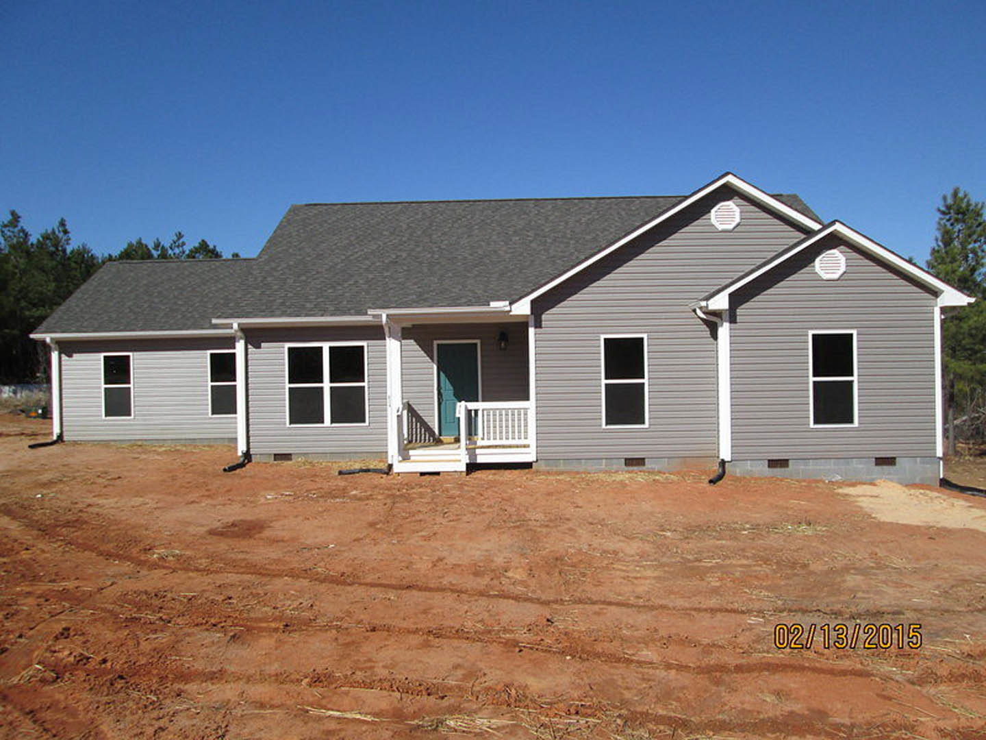 Grey siding house under construction with blue front door, white porch railing, and white fence, surrounded by dirt ground