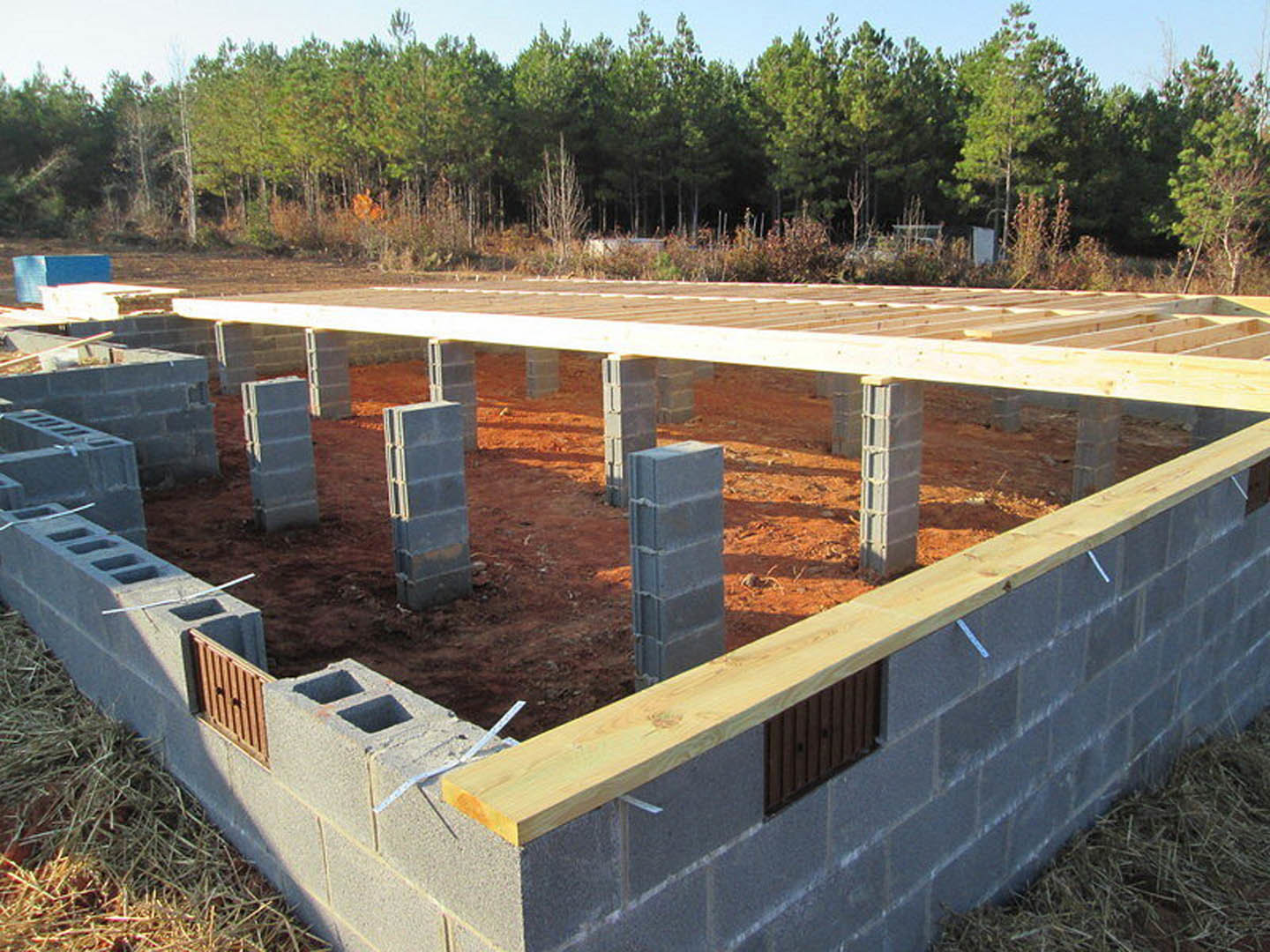 Wood-framed foundation under construction with concrete blocks, surrounded by trees and open sky