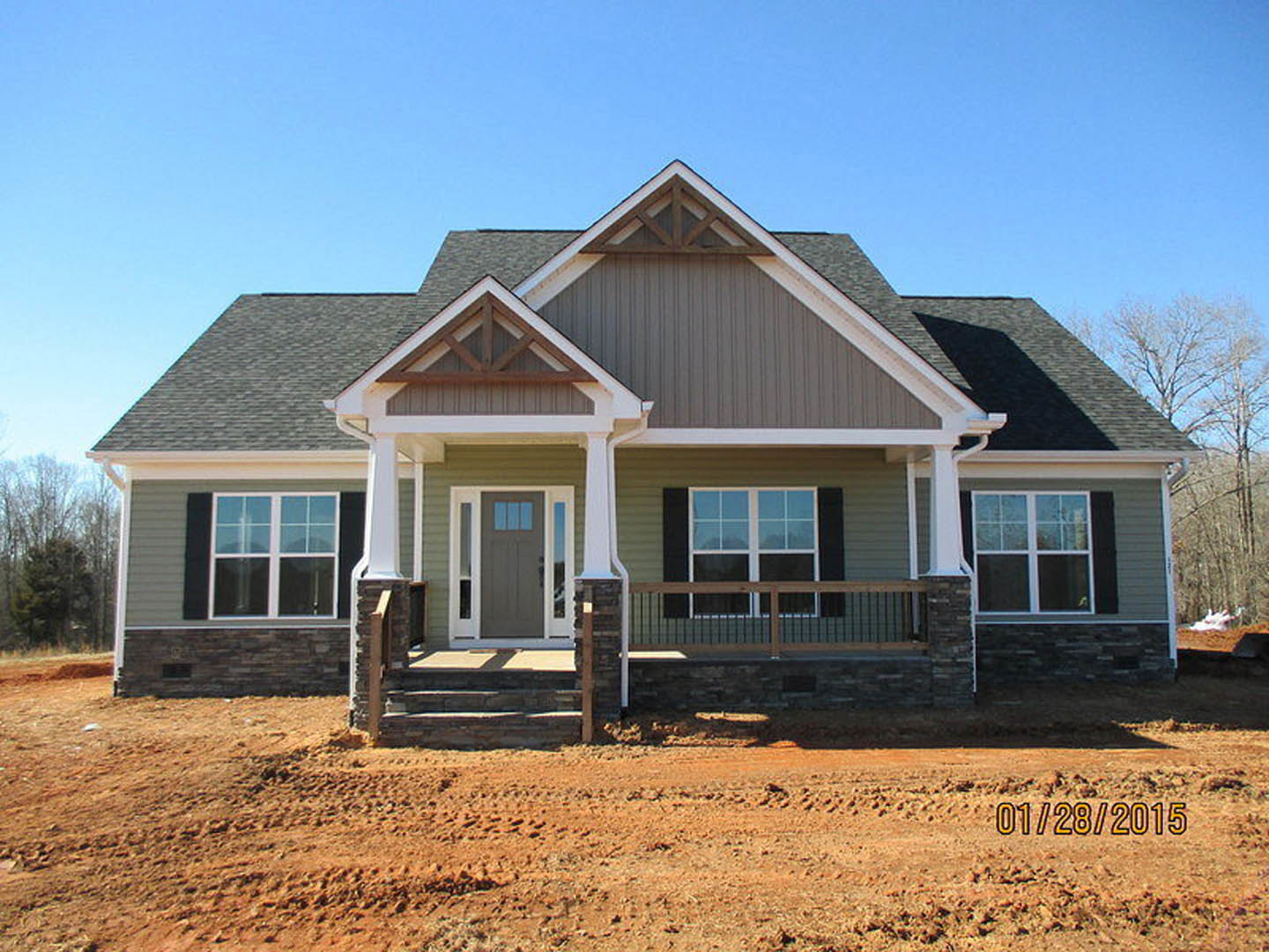Partially built house with white siding, covered porch, grey door with glass panels, white-framed windows, and dirt yard with a white pole