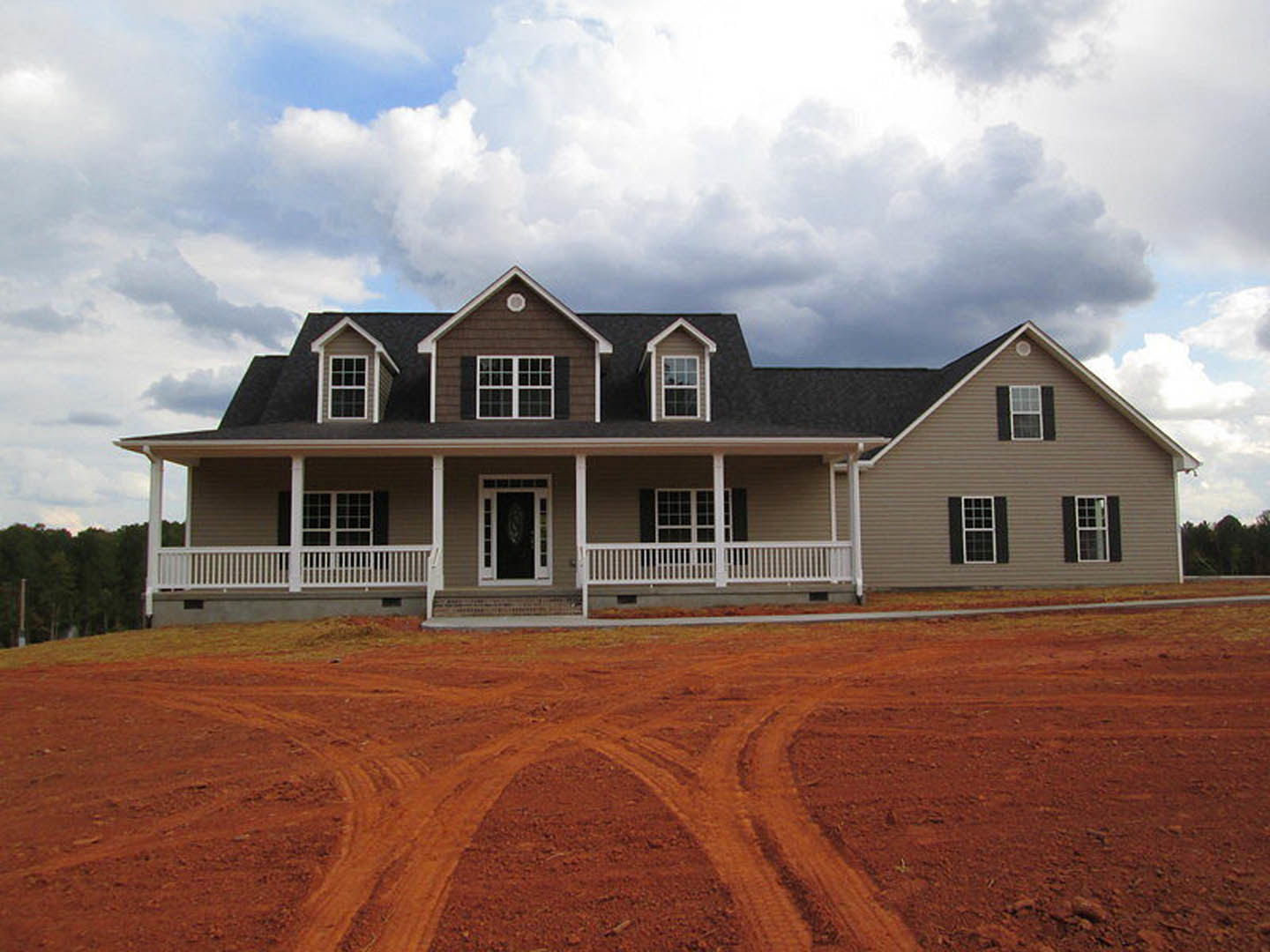 Two-story home with large gabled roof, multi-pane windows, covered front porch, and tire tracks on dirt driveway leading through open field.