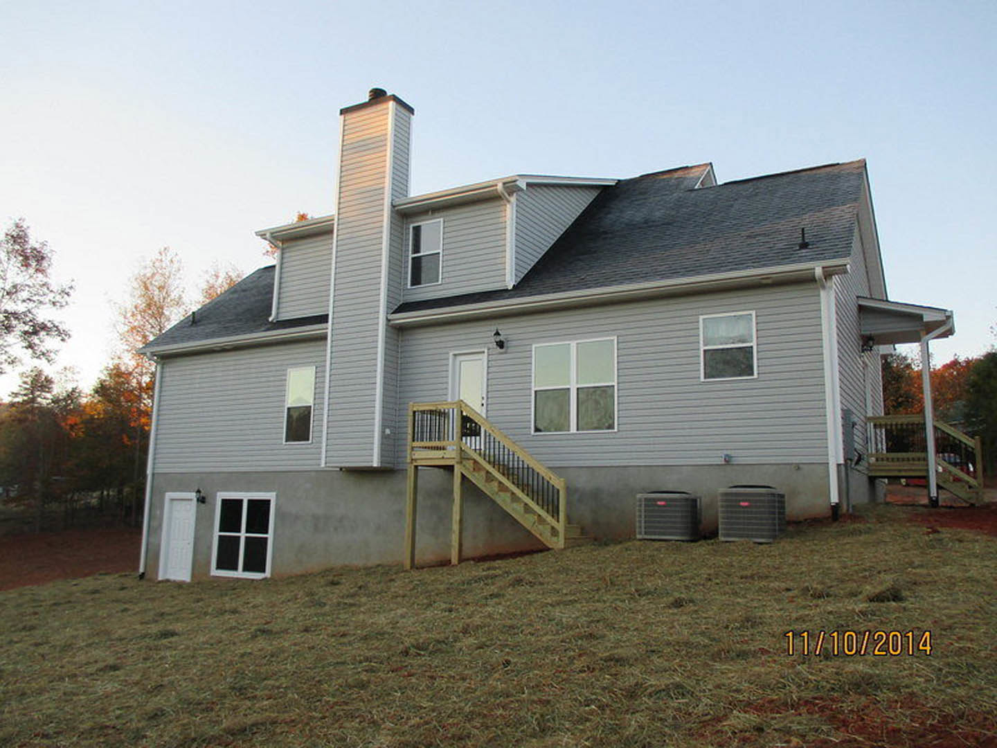 Two-story home with wood staircase leading to covered porch, large windows, and exterior air conditioning units; surrounded by trees under blue sky.