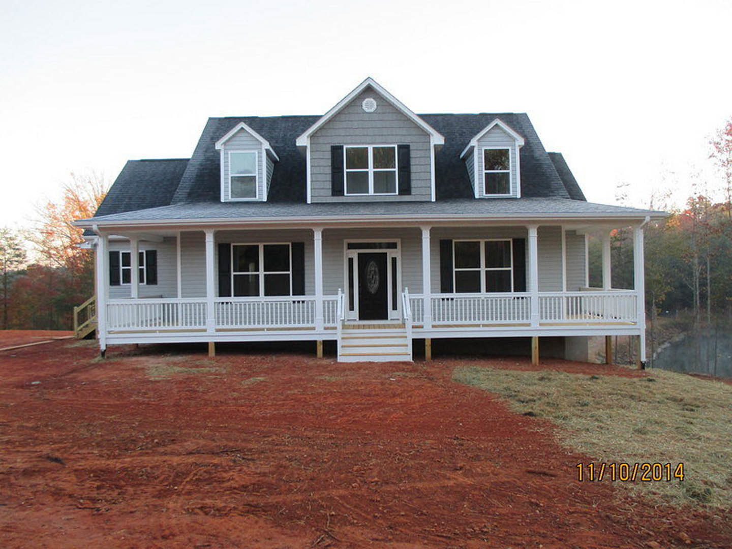 White house with covered porch, black door framed in white, white stairs and railing leading to entrance, red dirt yard in foreground, windows along exterior, tree visible to the