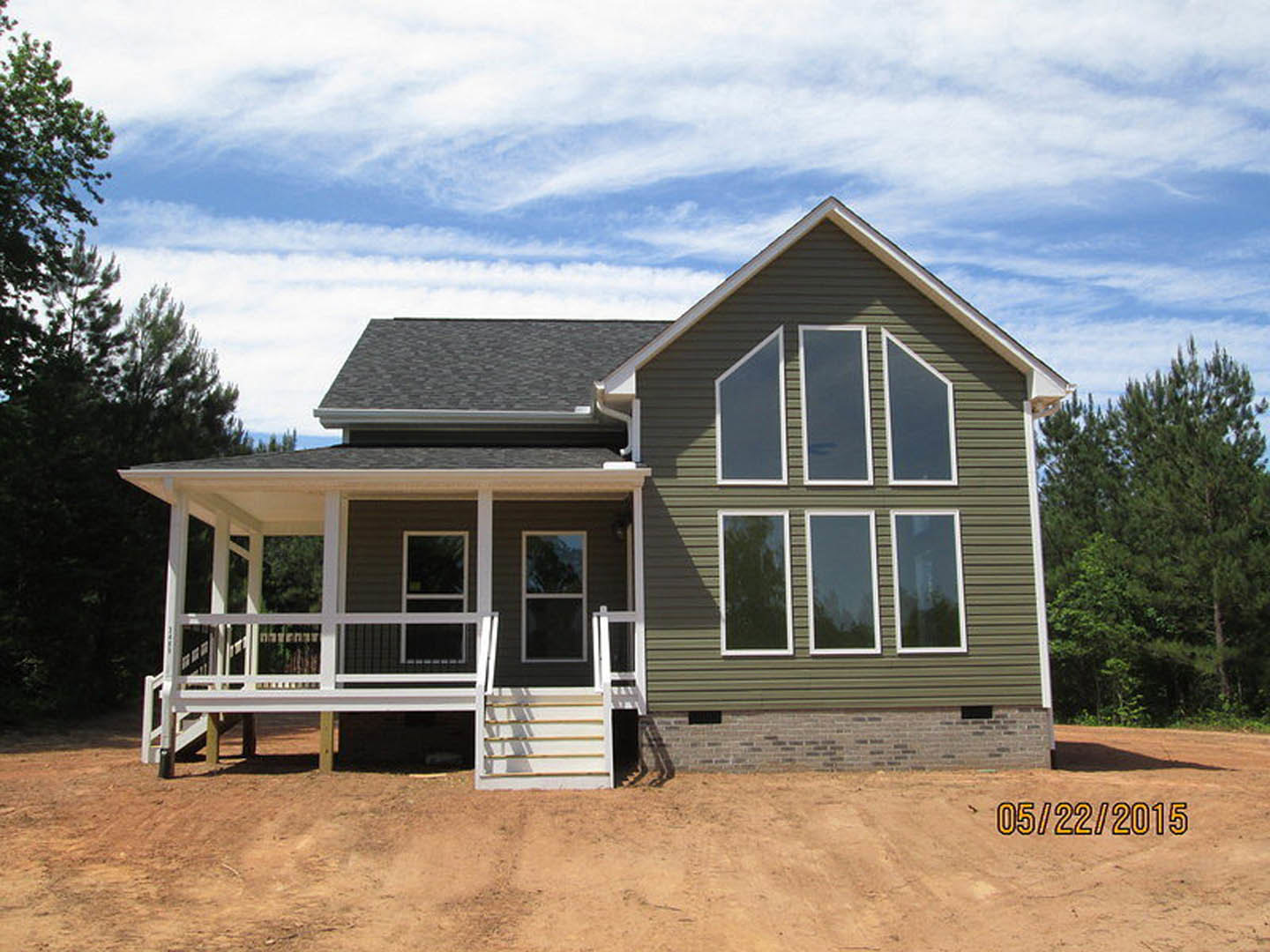 Two-story home with spacious front porch, white railings, multiple large windows with white frames, surrounded by mature trees under a partly cloudy sky