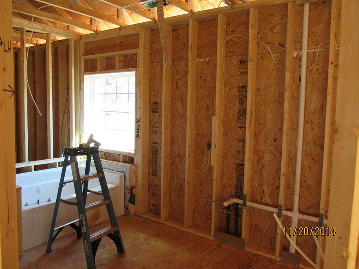 Bathroom under construction with exposed wooden beams, ladder leaning against unfinished bathtub, plywood flooring, and sunlight streaming through window
