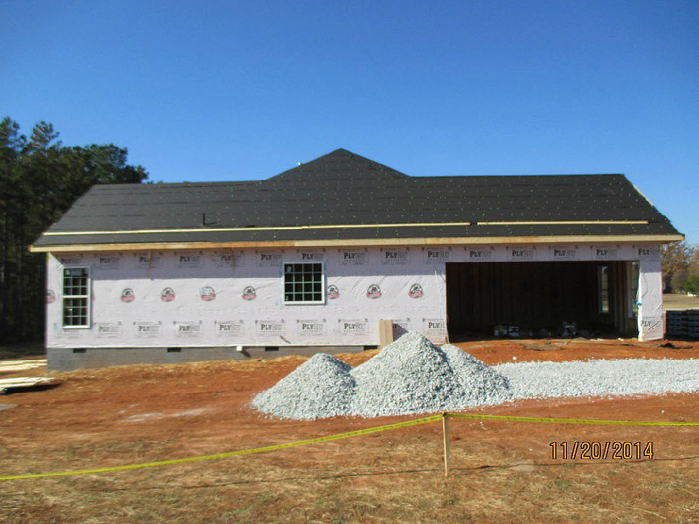 House under construction with black roof, white gravel pile in front, multi-pane window, yellow caution tape on dirt field, trees and sky in background