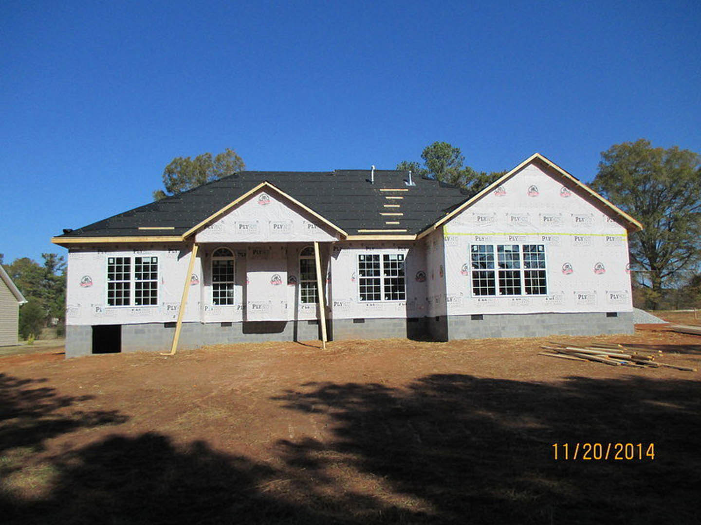 Framed house under construction with exposed wood, ladder leaning against siding, tree casting shadow on dirt, blue sky overhead