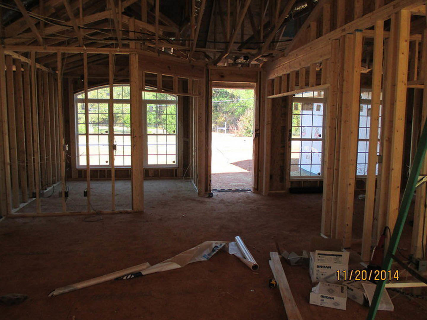 Open room with light wood flooring, exposed wooden beam on the ground, multiple windows including a large multi-pane window, door leading outside, view of wooden fence through