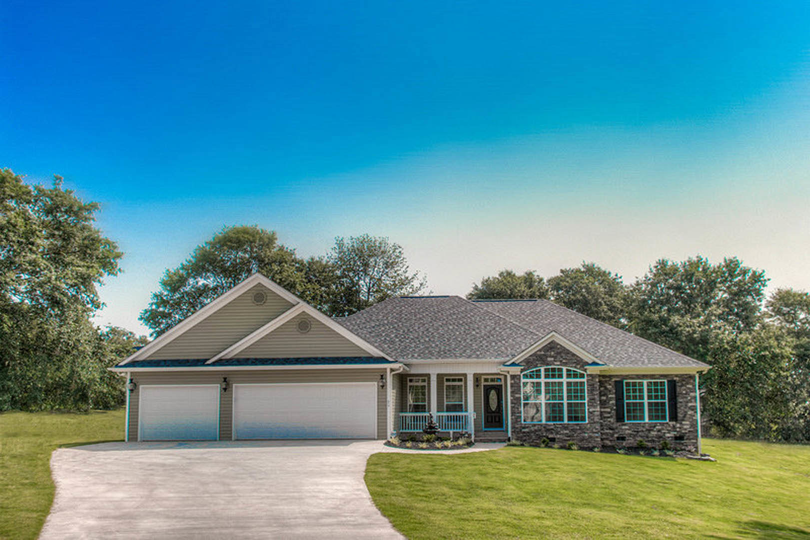 Two-story home with gray siding, white trim, attached garage, concrete driveway, manicured lawn, front porch, and mature trees in the background