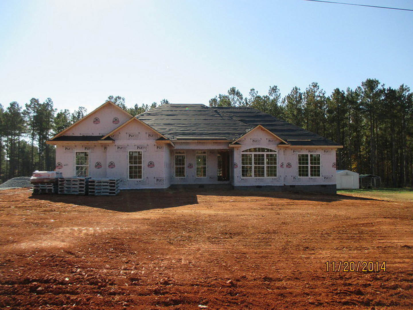 Partially built house with white exterior walls, black roof, grid-style windows, surrounded by dirt lot and mature trees in the background