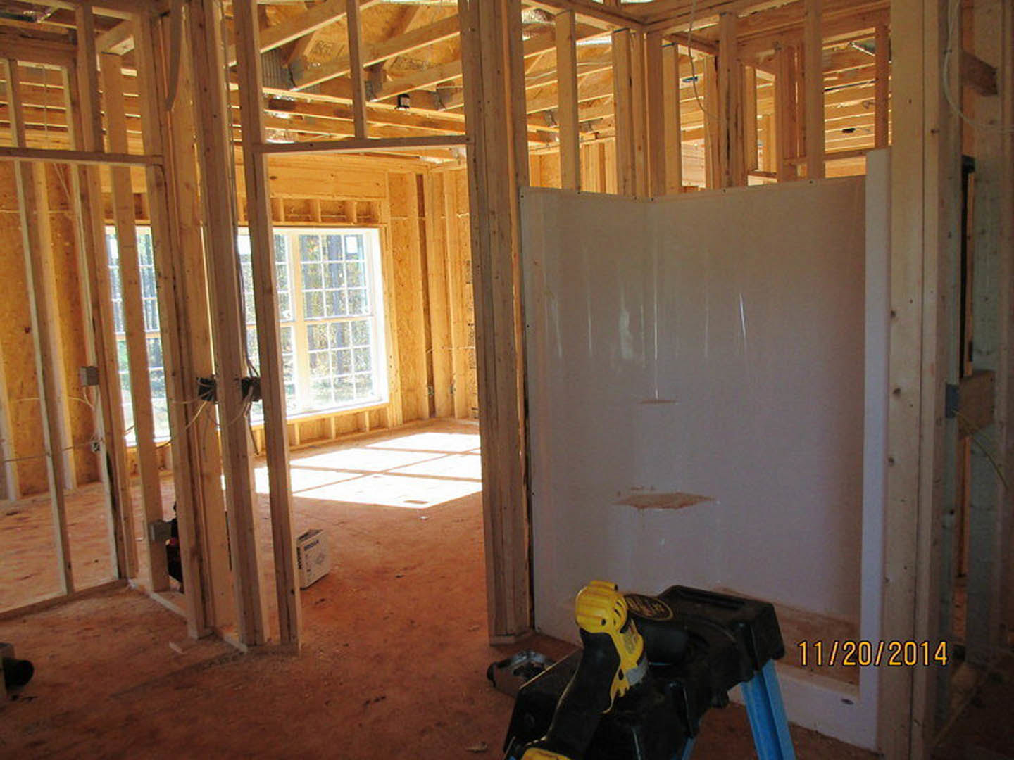 White wall with a wooden-framed window, yellow and black drill resting on a black container, yellow handle visible, construction materials and tools scattered in a partially
