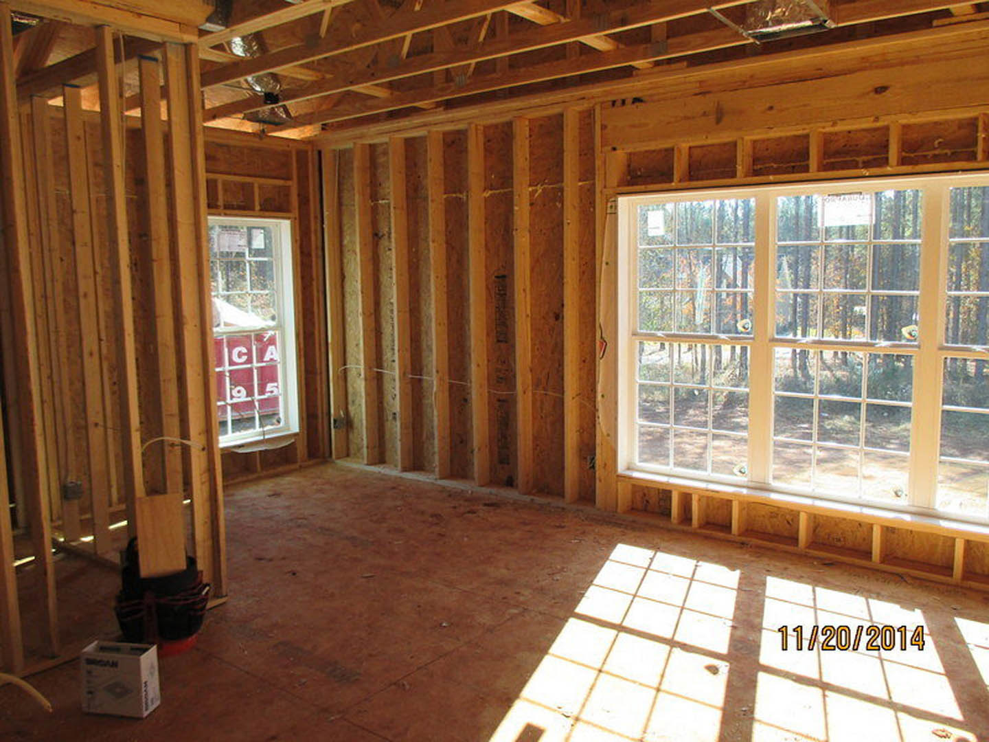 Sunlit room featuring a large window with natural wood framing, exposed wooden ceiling beams, light-colored walls, and hardwood flooring.