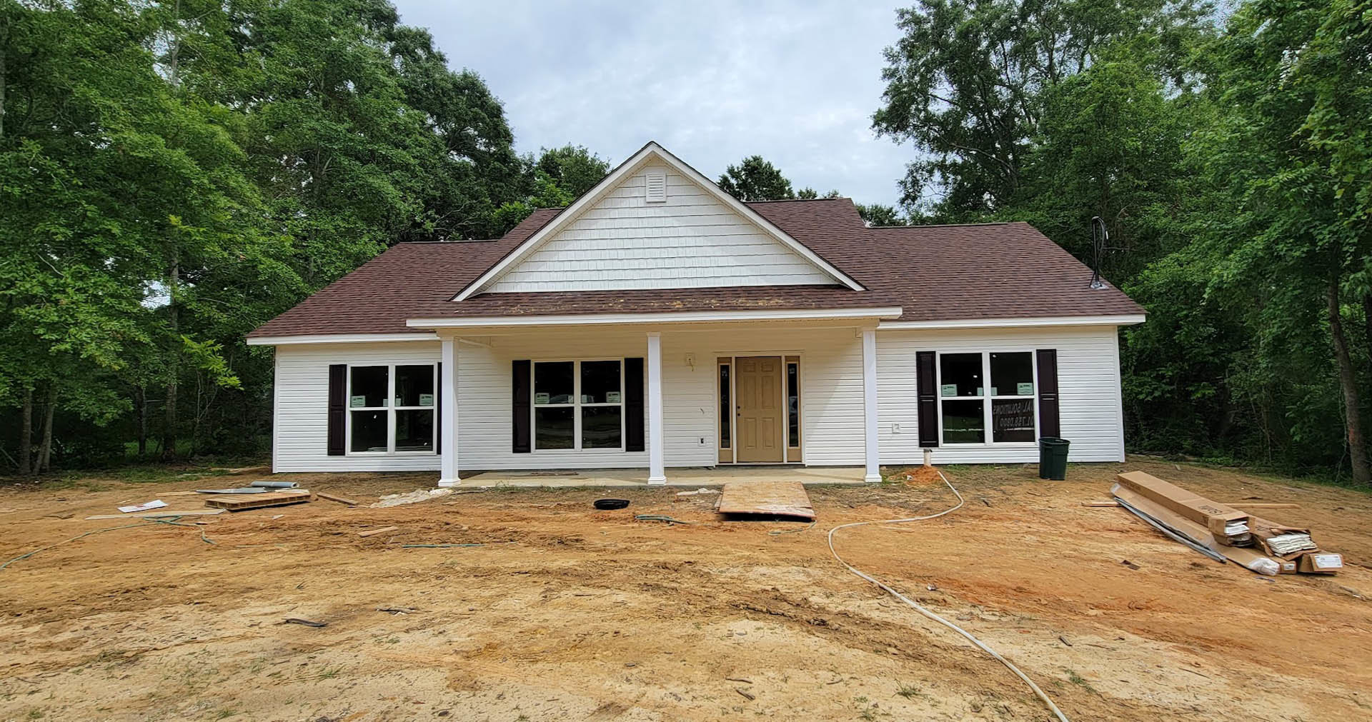 Framed house under construction with exposed plywood walls, white-trimmed window, porch area, and trees in the background