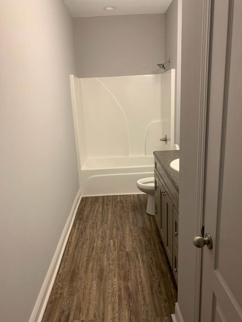 Bathroom with white sink and toilet, wood laminate flooring, wall-mounted shower head, white plaster walls, close-up of chrome door knob