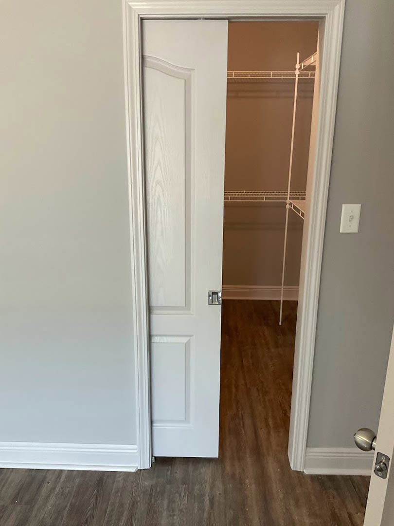 Open closet with wood flooring, white walls and trim, white door with silver knob, visible wall outlet and light switch