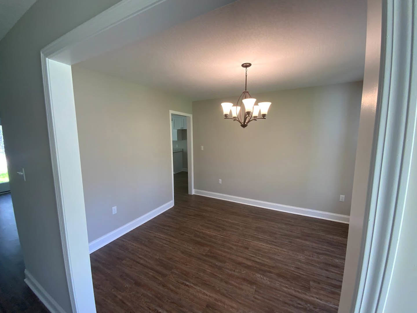 Wood flooring with white baseboards, glass-paneled door, ornate chandelier hanging from plaster ceiling, hallway visible through open doorway.