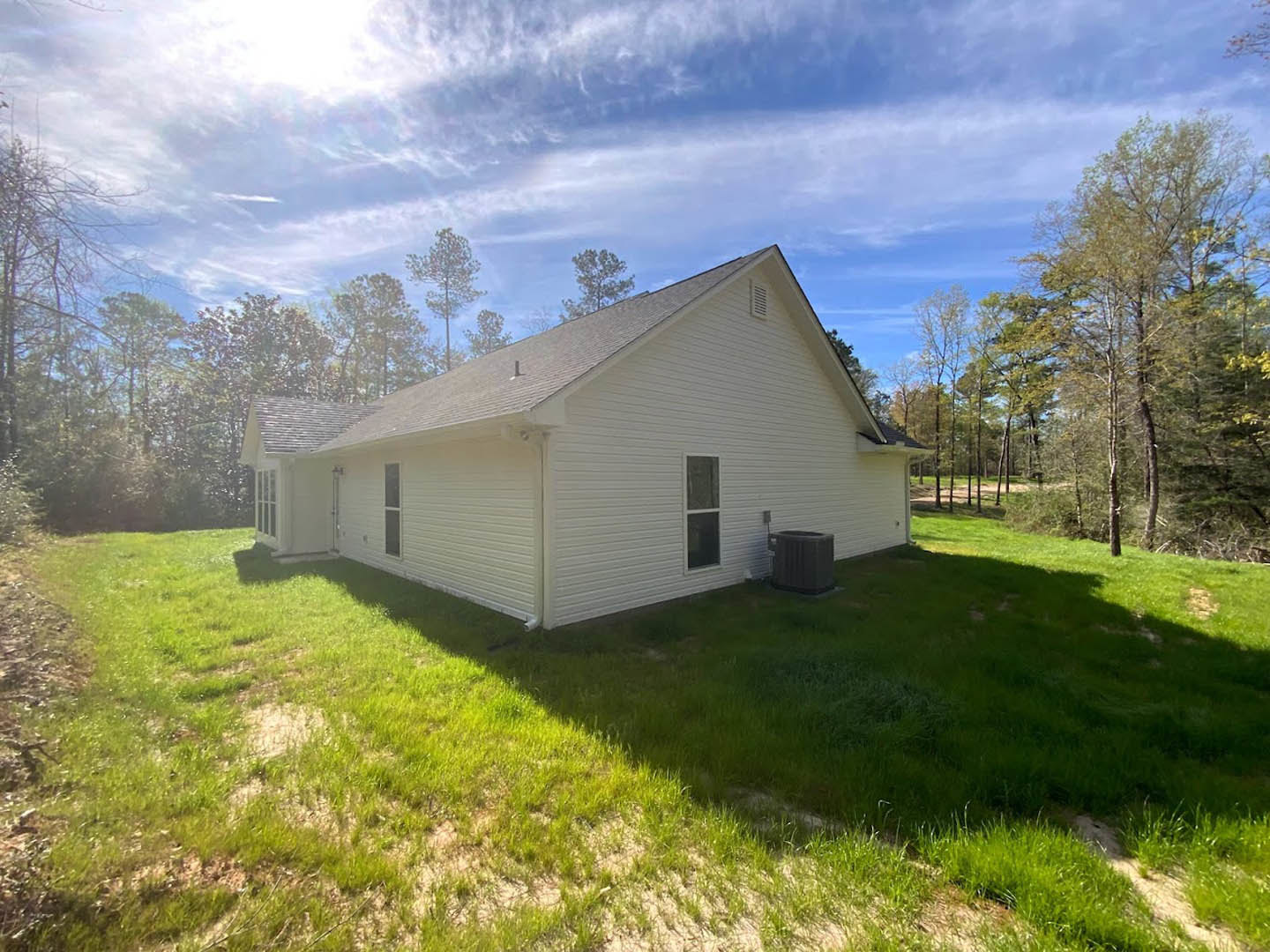 White house with large windows, green lawn in front, black trash can near entry, mature trees in background, blue sky with scattered clouds