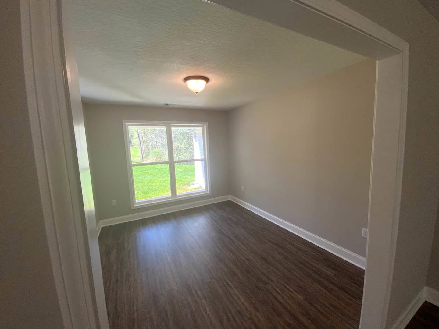 Room with wood flooring, white walls, large window overlooking green grass, ceiling light fixture, simple molding along baseboards