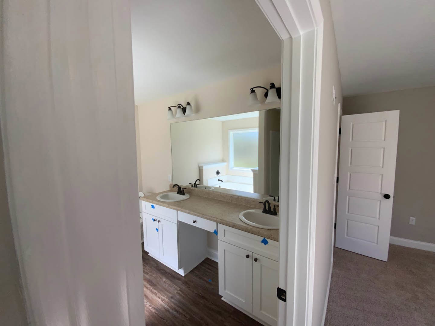 Bathroom with double vanity featuring white sinks, black faucets, large framed mirror, white cabinetry, light gray tile floor, and a white door with black handle