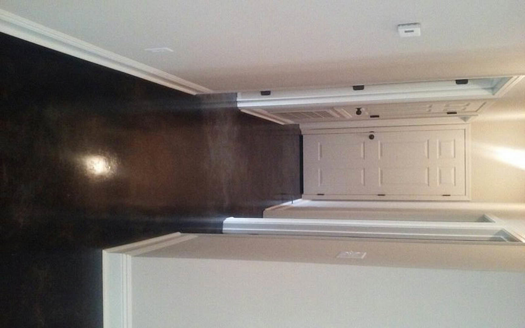 Hallway with white walls and ceiling, white door featuring a round knob, silver-framed accent, black object mounted on ceiling, white box with black stripe, cabinetry visible in