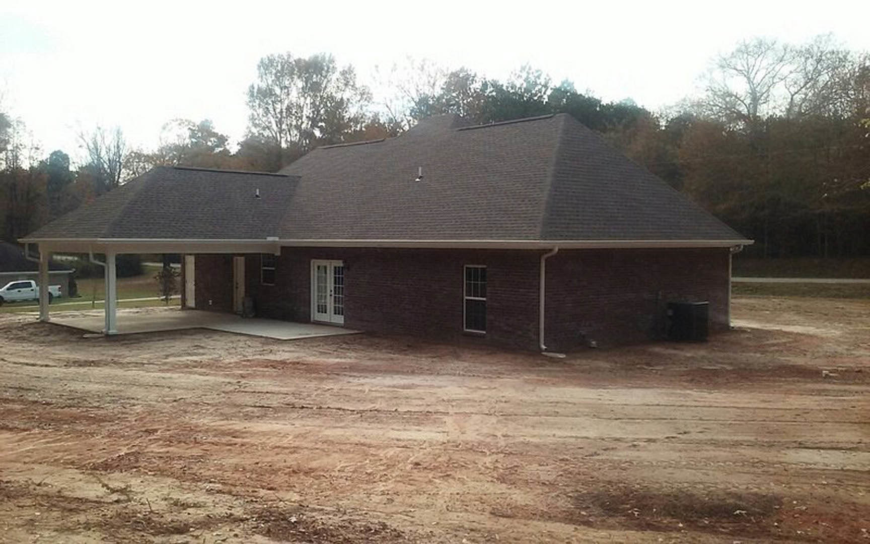 Partially built house with exposed framing, shingled roof, glass pane door, large windows, dirt yard, white pickup truck, surrounding trees.