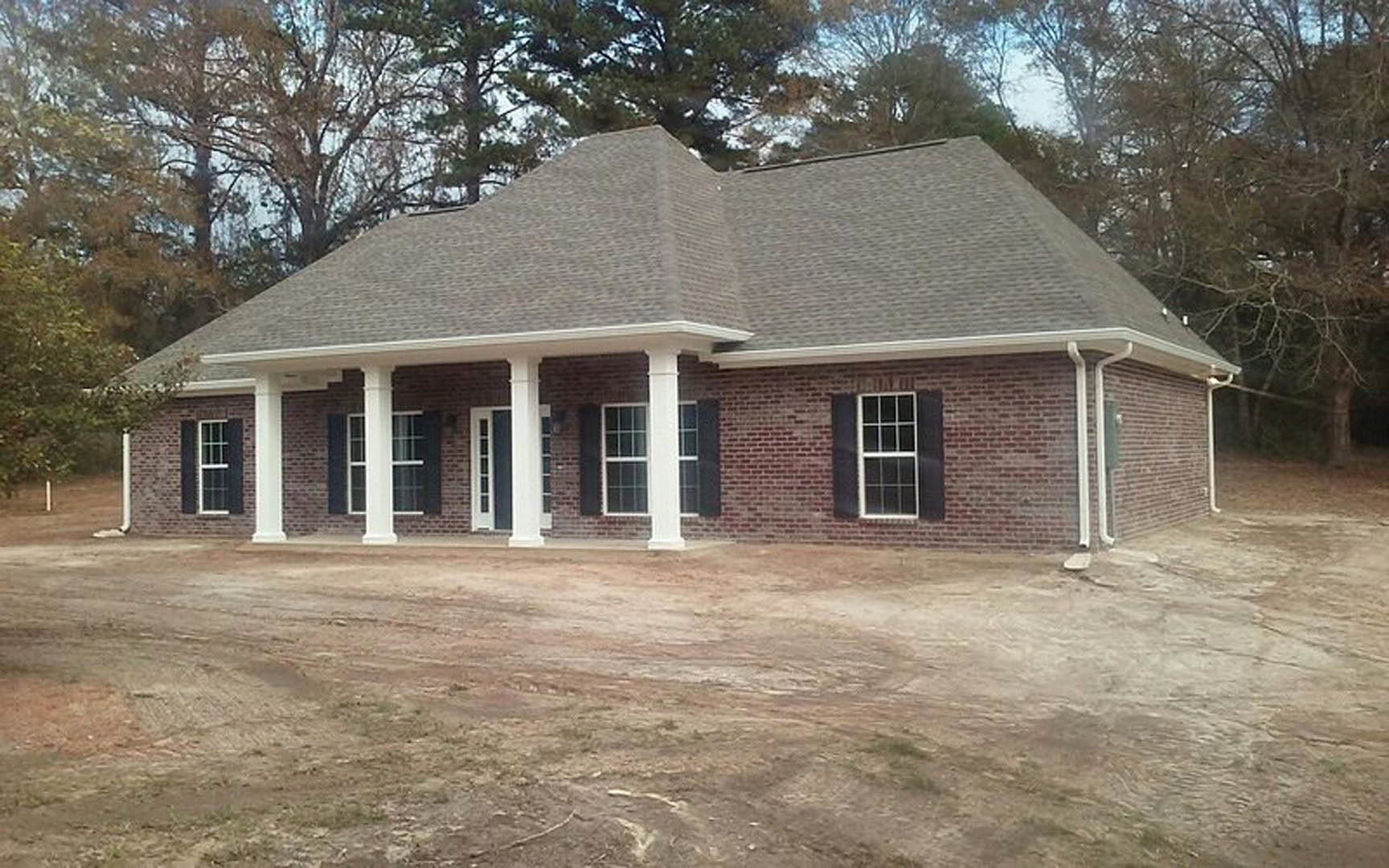 Red brick house with white columns, white-framed windows featuring grid patterns, surrounded by trees and landscaping under a blue sky