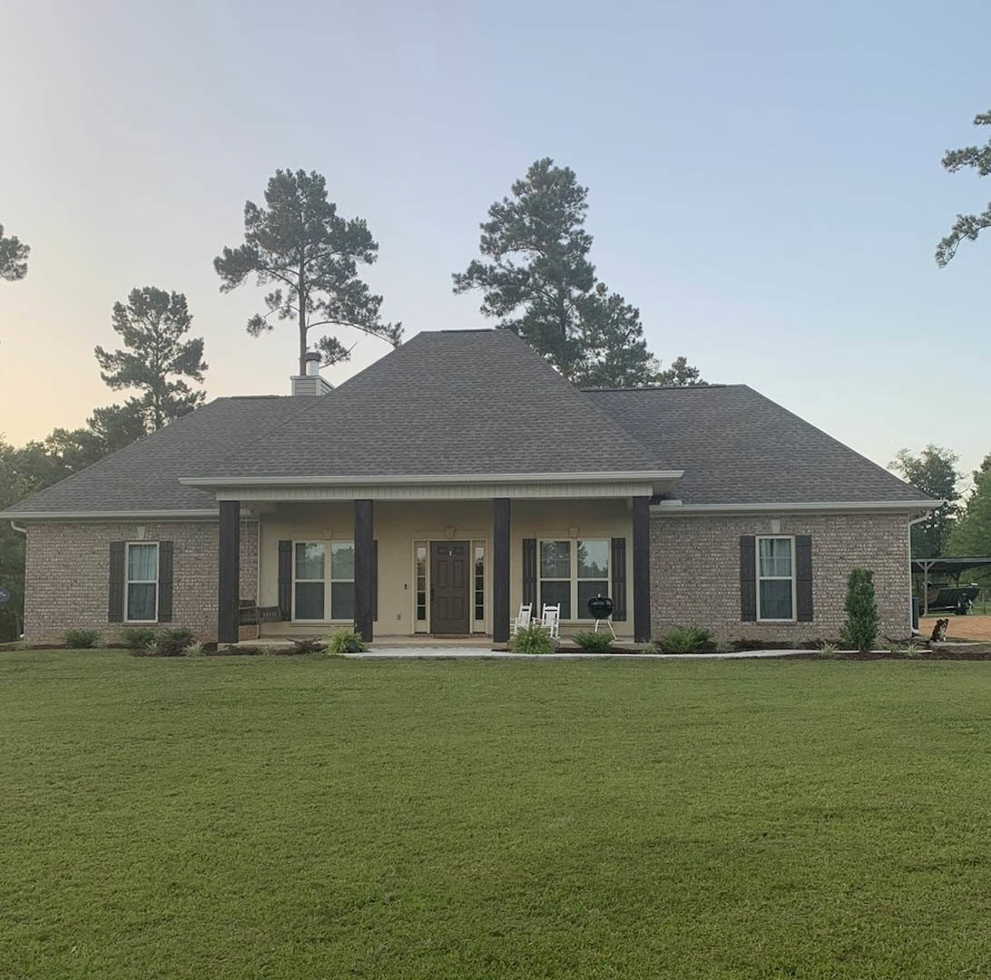 Two-story farmhouse with white siding, covered porch featuring a grill, manicured green lawn, tall tree, street light, white-framed windows, and blue sky in the background