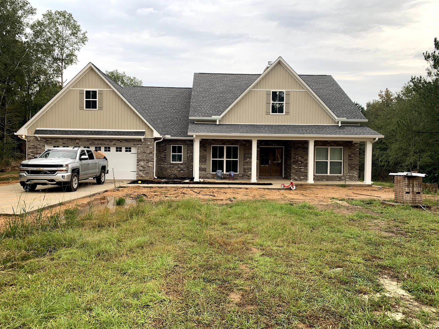 Silver pickup truck parked on driveway in front of modern house with double glass doors, white trim, and large windows; manicured lawn and trees in background.