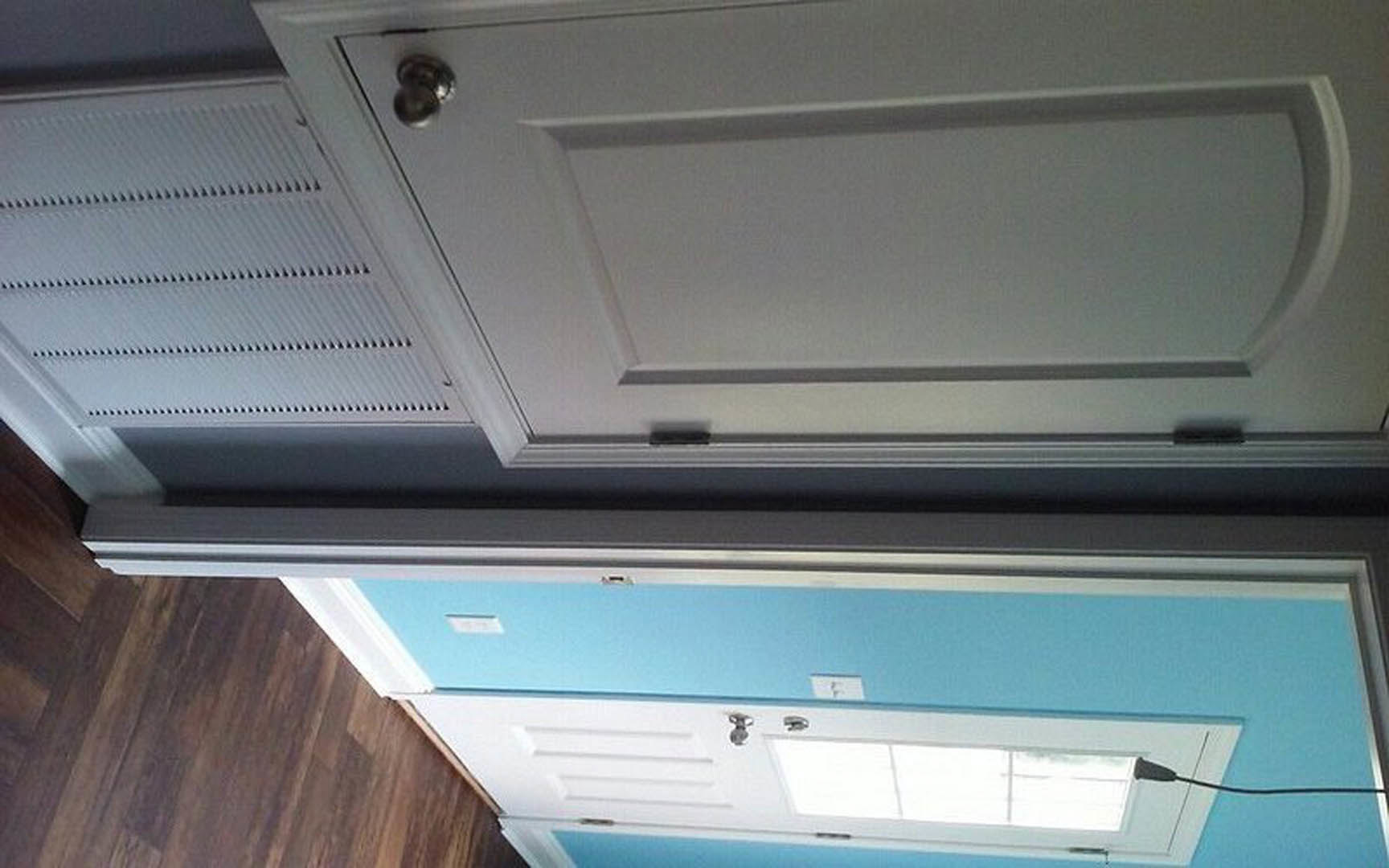 White interior door beside a white cabinet with silver handle, wall-mounted vent, and window showing daylight sky.