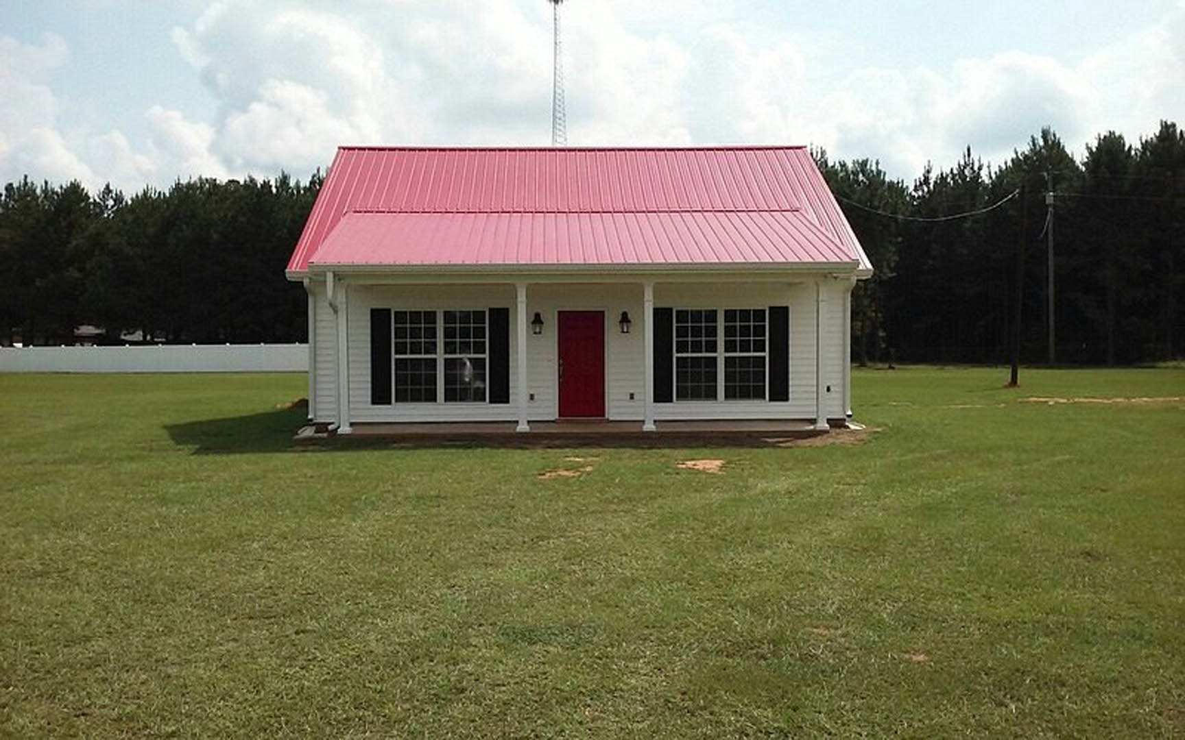 Two-story home with red metal roof, white siding, red front door with white trim, multiple square windows, green lawn, trees, and cloudy sky; radio tower visible in background