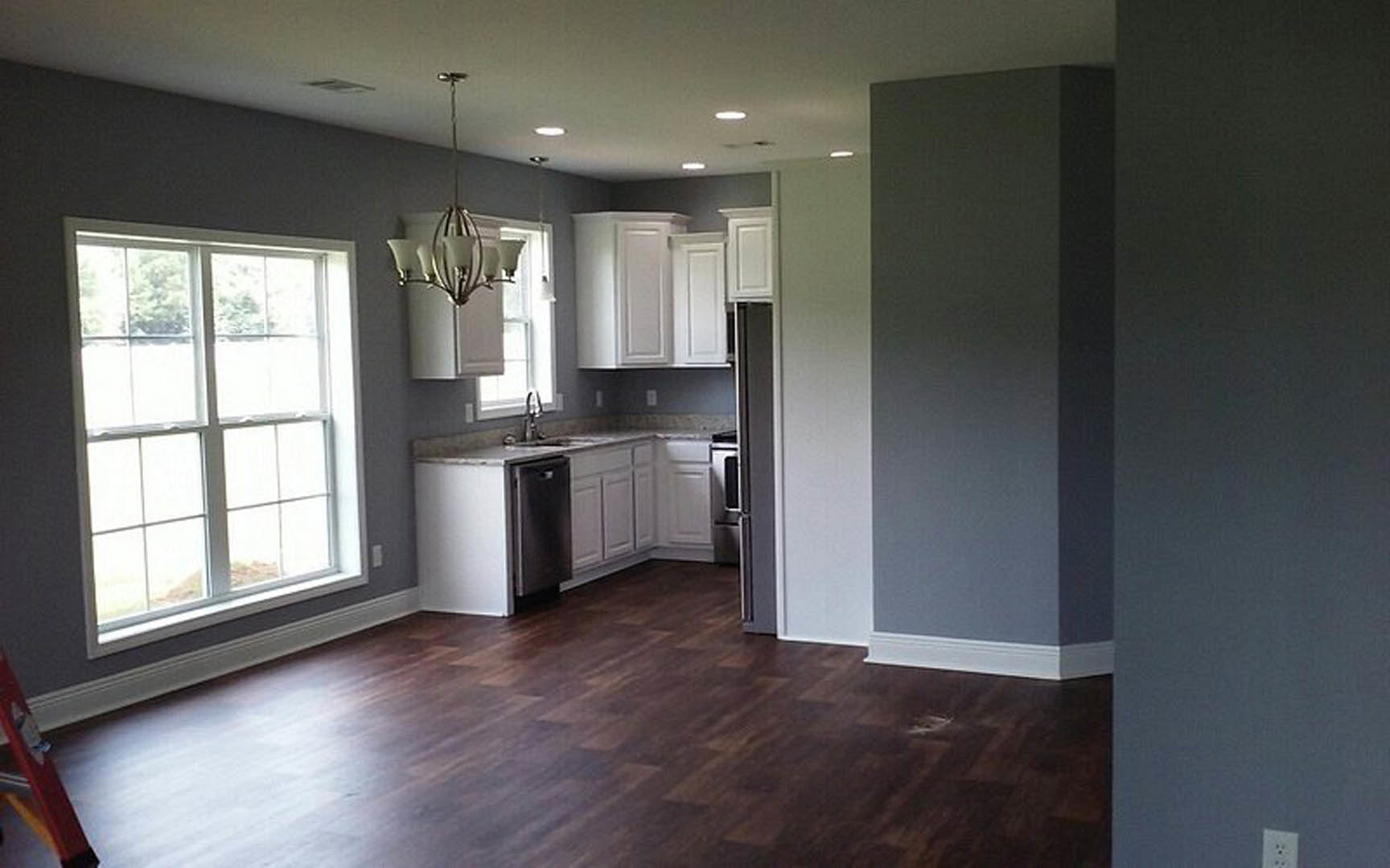 Kitchen with white shaker cabinets, dark wood flooring, multi-pane window, and a chandelier hanging above the space; red ladder visible in the background.