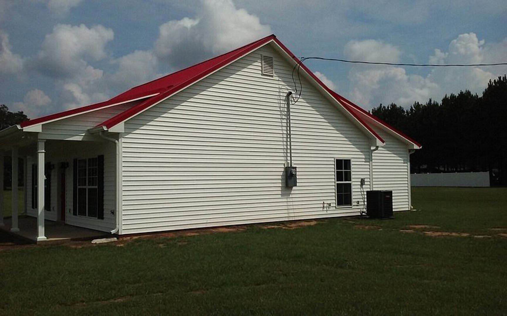 White siding house with red gabled roof, white framed windows with cross muntins, grassy lawn, exterior vent, black electrical fixture attached to wall, cloudy sky and trees in