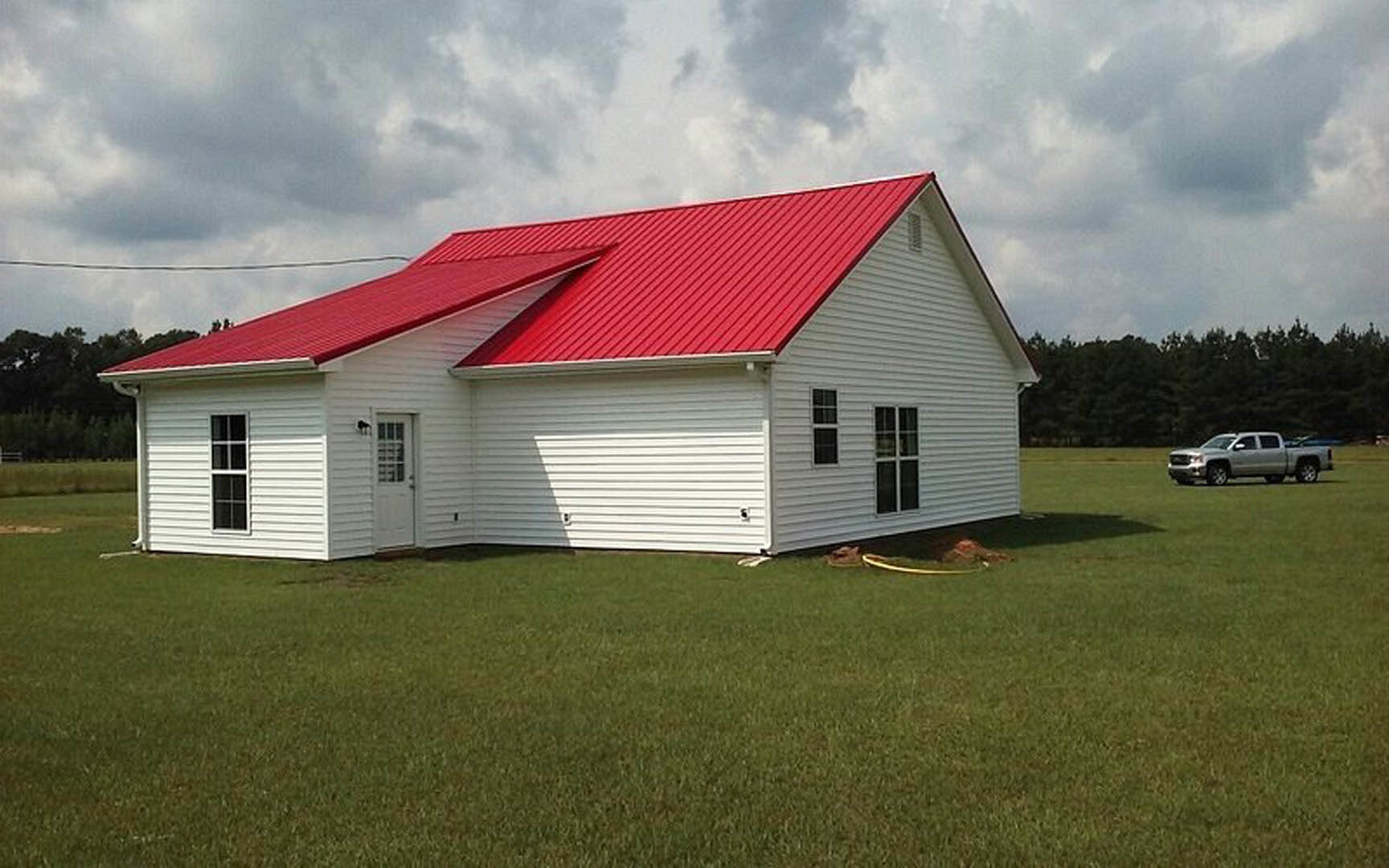 White farmhouse with red gabled roof, white framed windows, white door with window, green lawn with yellow hose, trees and cloudy sky in background
