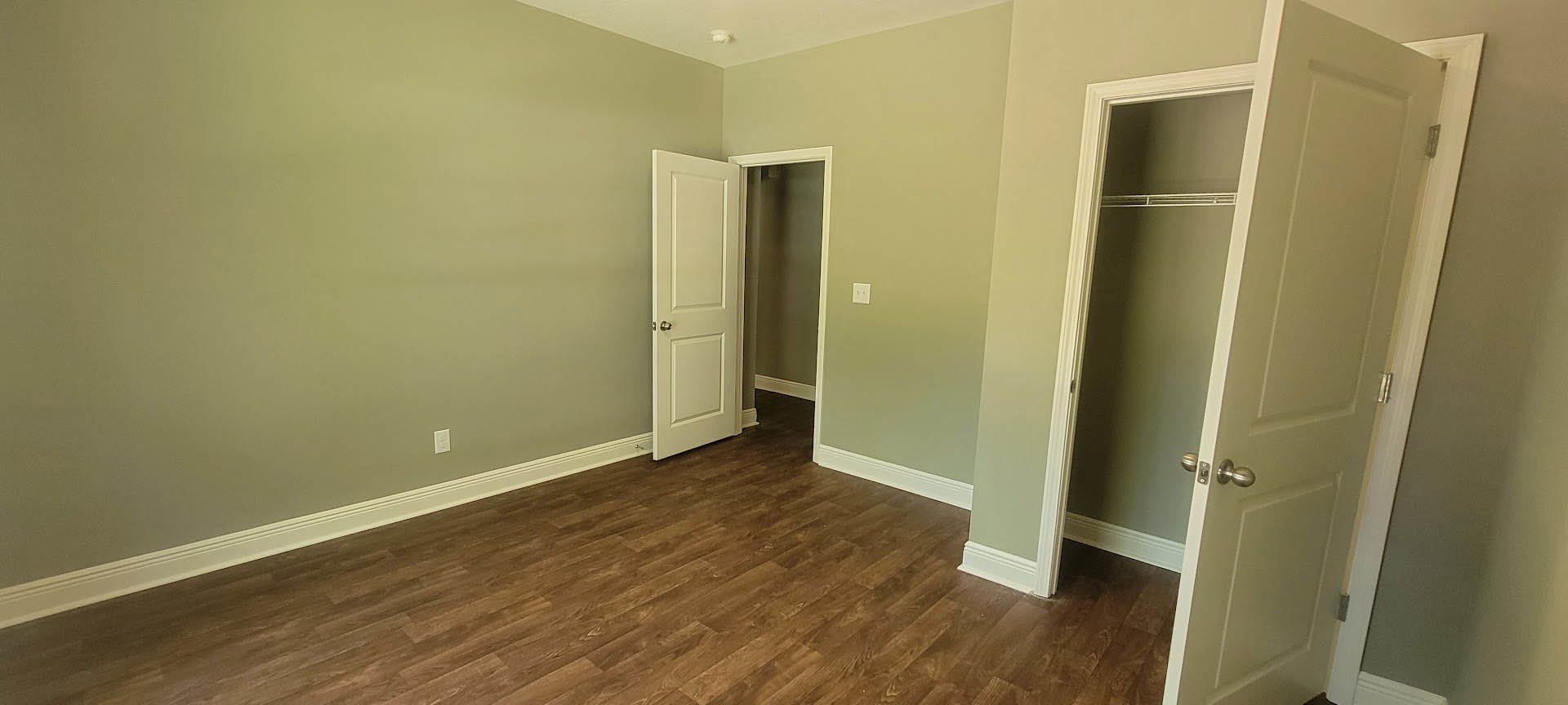 White-painted room with wood laminate flooring, two doors open revealing a closet and bathroom, both doors featuring silver knobs and white trim.