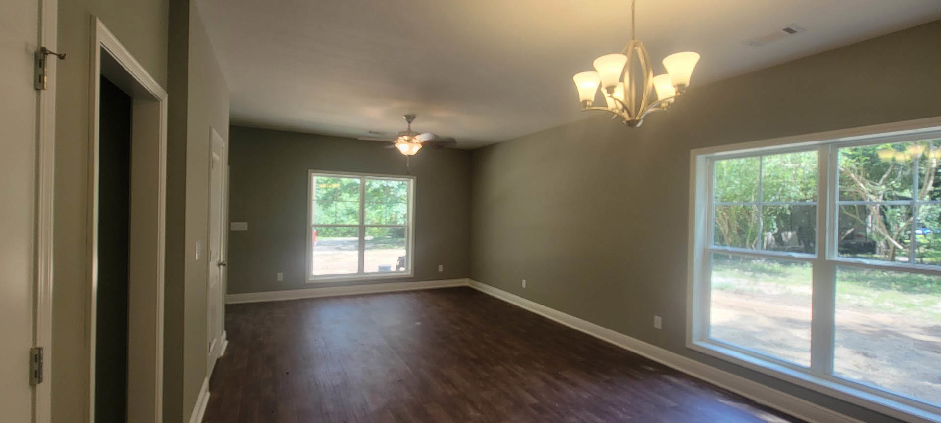 Room with dark wood flooring, white walls, ceiling fan, large window showing trees outside, decorative chandelier, crown molding