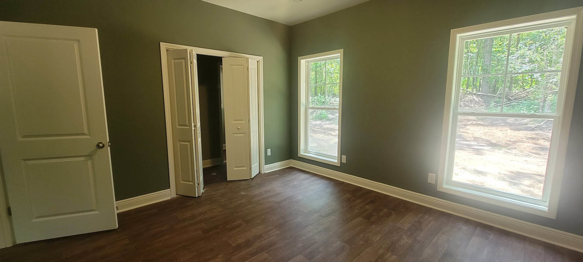 Open white door with silver handle leading to room with wood flooring, large window showing forest view, white walls, and natural light.