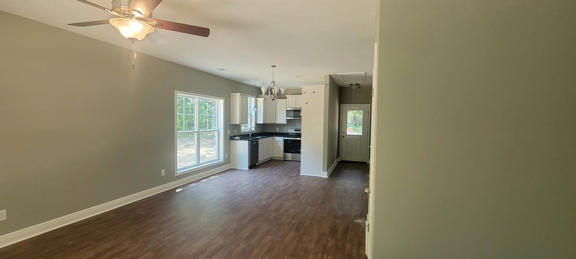 Open-concept kitchen and living room featuring wood flooring, white cabinetry with shelving, ceiling fan with light fixture, large window offering outdoor views, and a white door