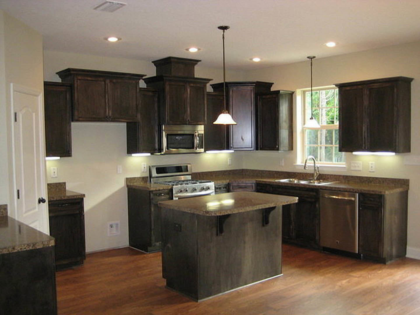 Kitchen with dark wood cabinets, marble-topped island, stainless steel microwave and stove, vent hood, and white square sink with faucet