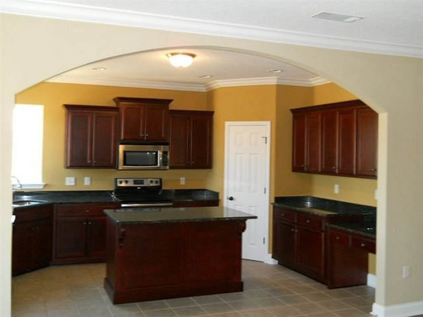 Kitchen featuring dark wood cabinetry, black countertop, stainless sink, white door with black handle, brown wooden surfaces, and blurred oven and light fixtures