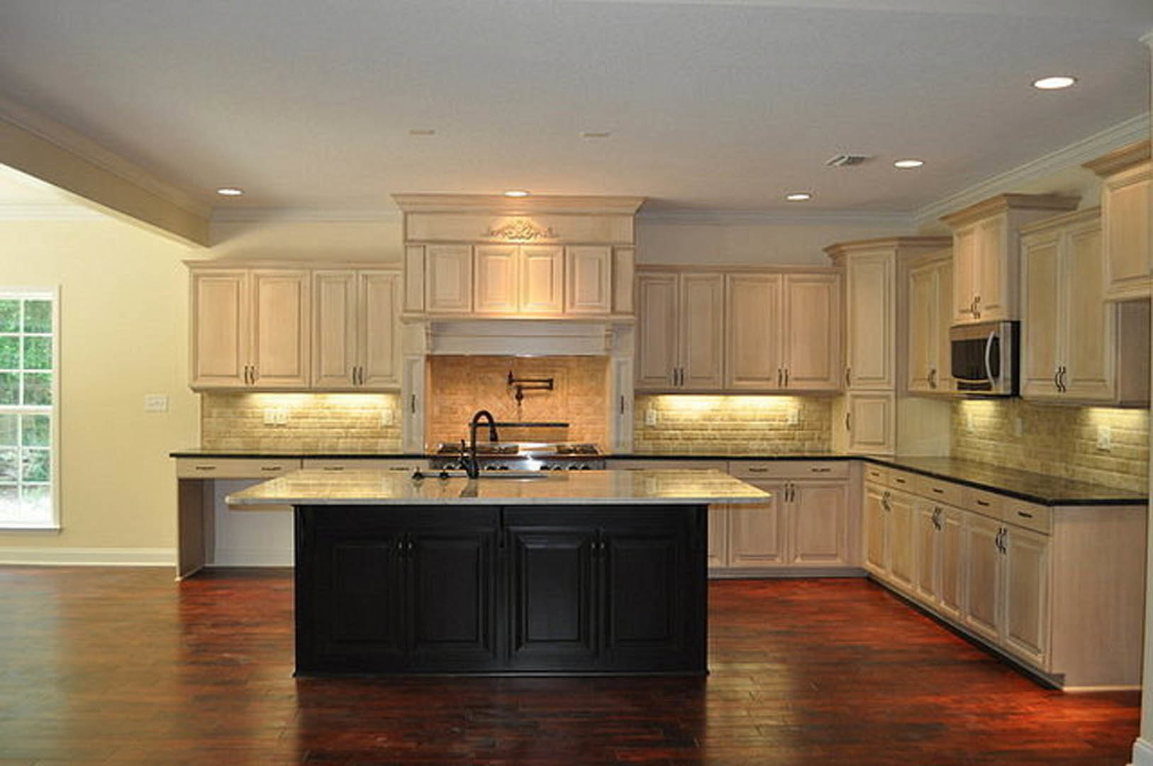 Spacious kitchen featuring a large granite island, dark wood flooring, black cabinetry, stainless steel microwave, tile backsplash, and a window above a sink