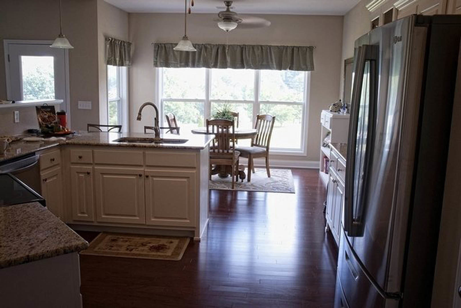 Modern kitchen with wood cabinetry, stainless steel refrigerator, dining table with upholstered chairs, potted plant on the countertop, large window, and light-colored flooring