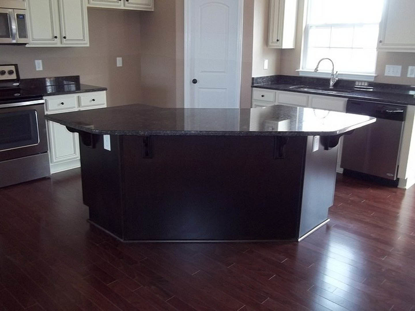 Spacious kitchen featuring a large central island with stone countertop, stainless steel appliances including a microwave and stove, white cabinetry, window above sink with modern