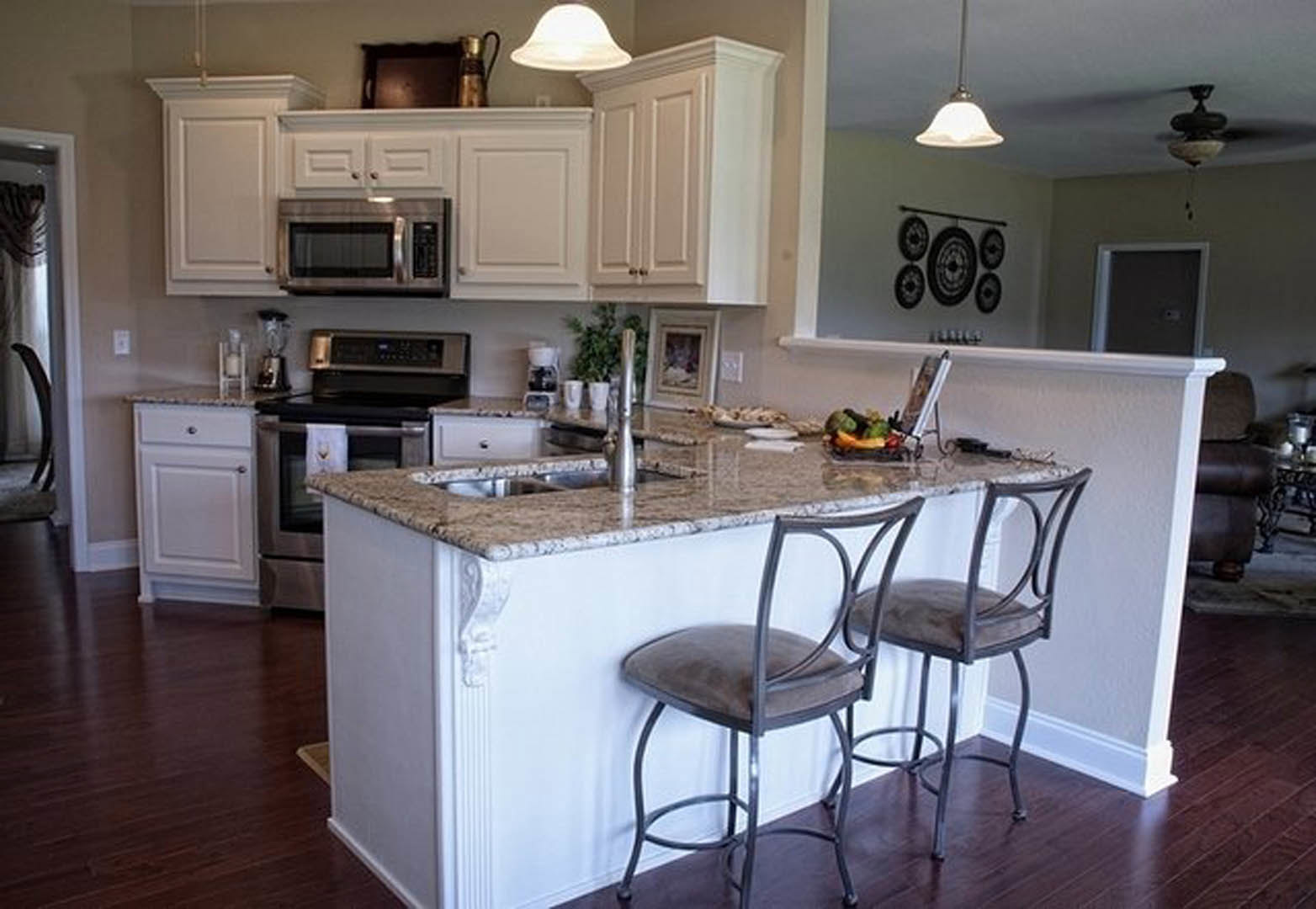 Modern kitchen with bar seating, mirrored backsplash, light cabinetry, stone countertops, stainless steel microwave, and two chairs placed against the wall