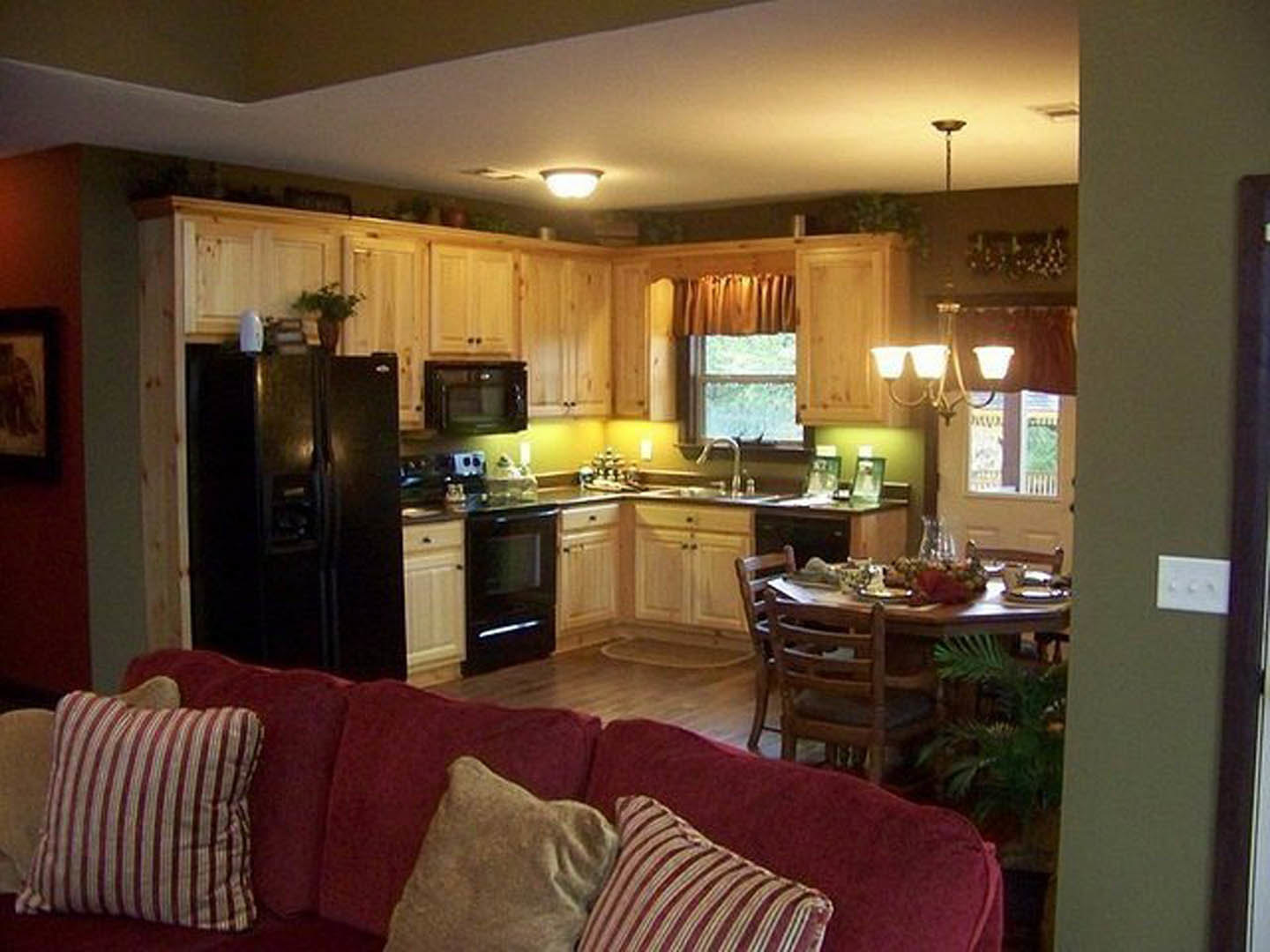 Open-concept kitchen and living room featuring wood cabinetry, black refrigerator, gray upholstered couch, dining table, potted plant, and large windows with natural light