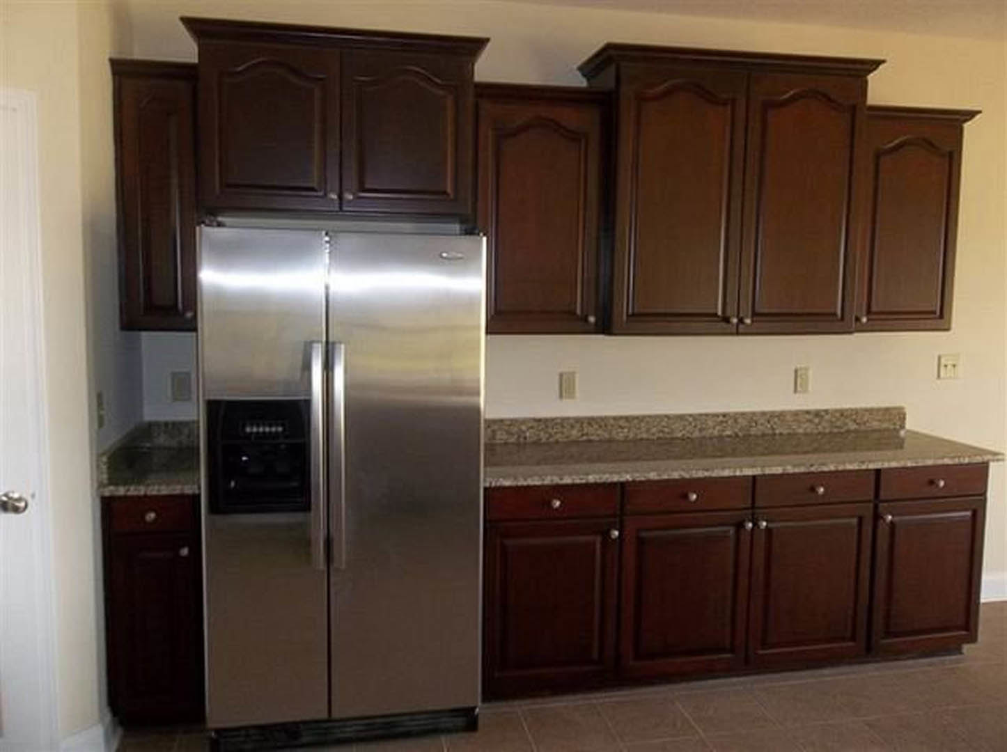 Stainless steel French door refrigerator beside dark wood cabinets with brushed metal knobs, granite countertop, and built-in oven in a modern kitchen