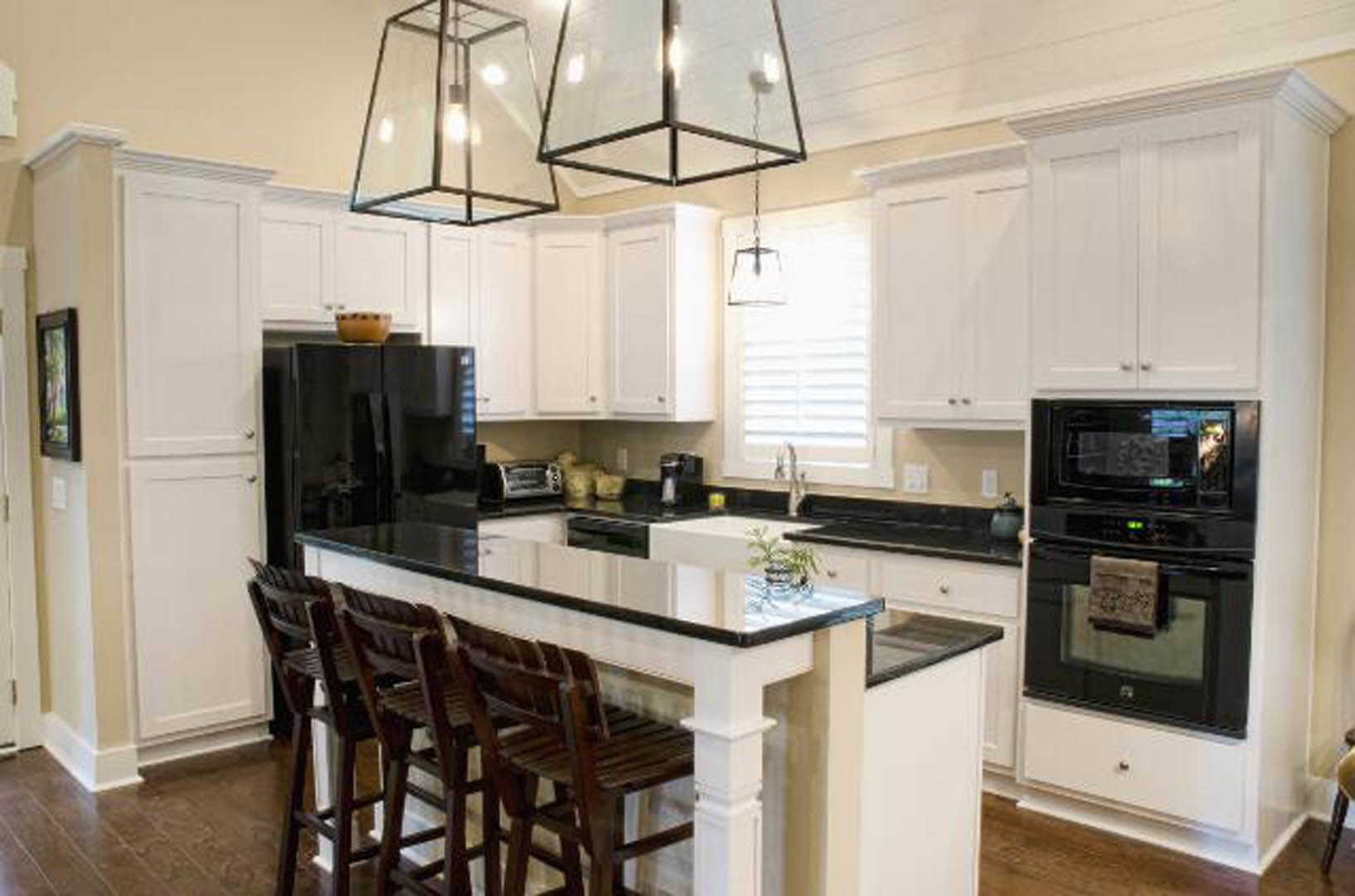 White kitchen cabinets with black countertops, black refrigerator and microwave, row of barstools at peninsula, modern pendant light fixture, neutral walls and window providing