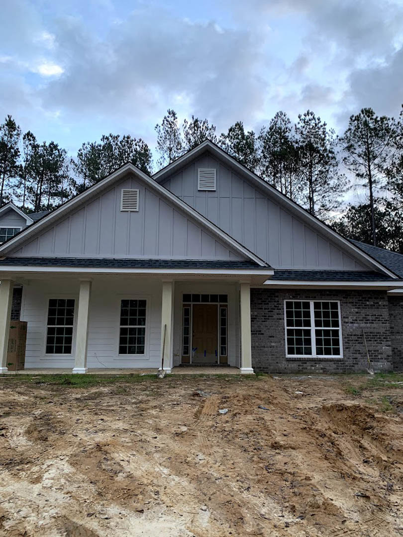 Wood-framed house under construction with multi-pane windows, glass-paneled door, covered porch, dirt ground with tire tracks, and trees in the background