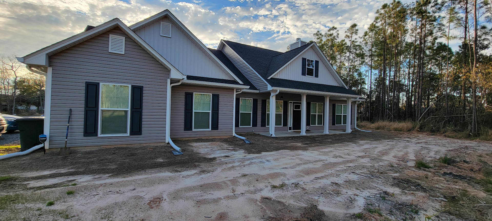 Two-story house with white siding, black shuttered windows, and a vent above the entry; dirt road and patch in front, surrounded by trees under a cloudy sky.