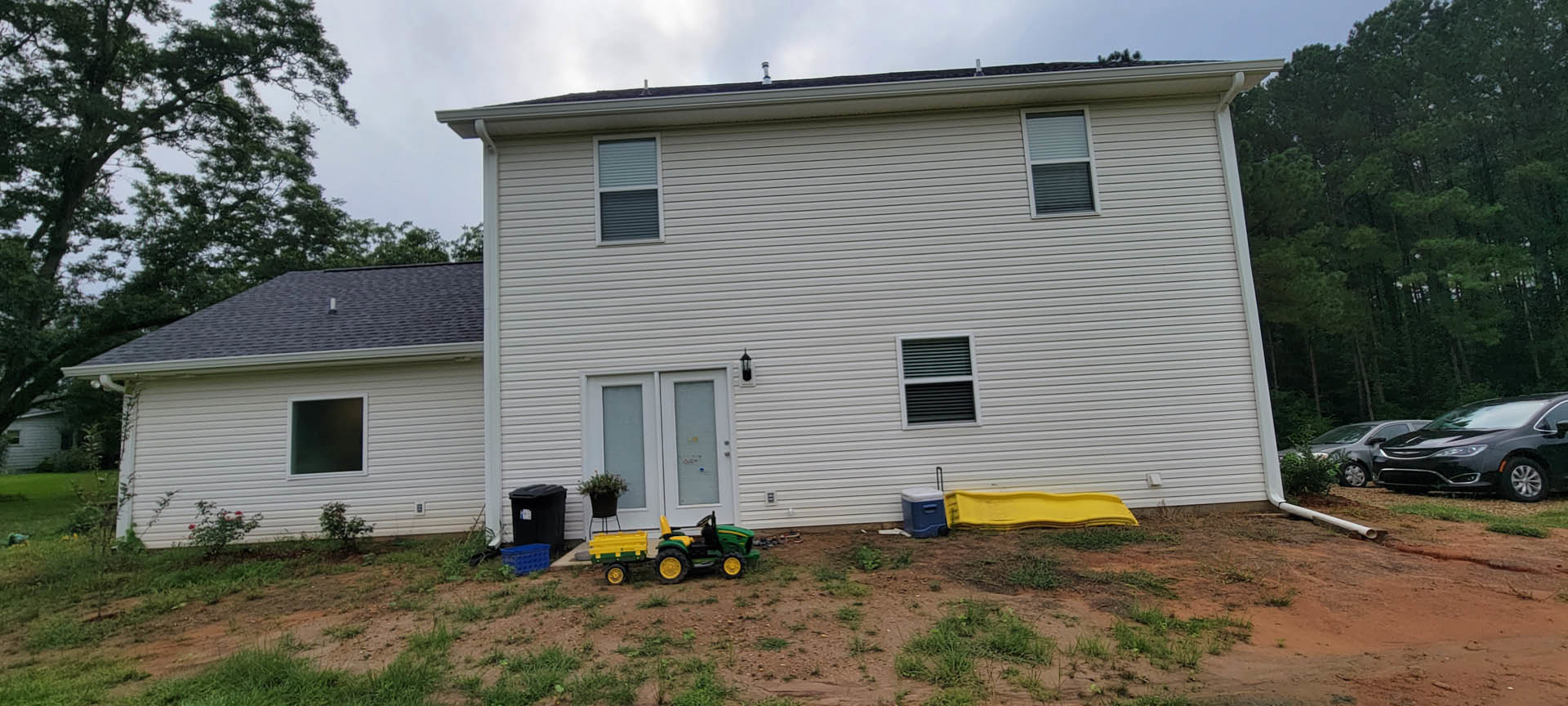 Two-story house with white siding, black roof, and large windows, surrounded by green lawn; yellow slide attached to play structure, lawnmower parked near driveway, black car in