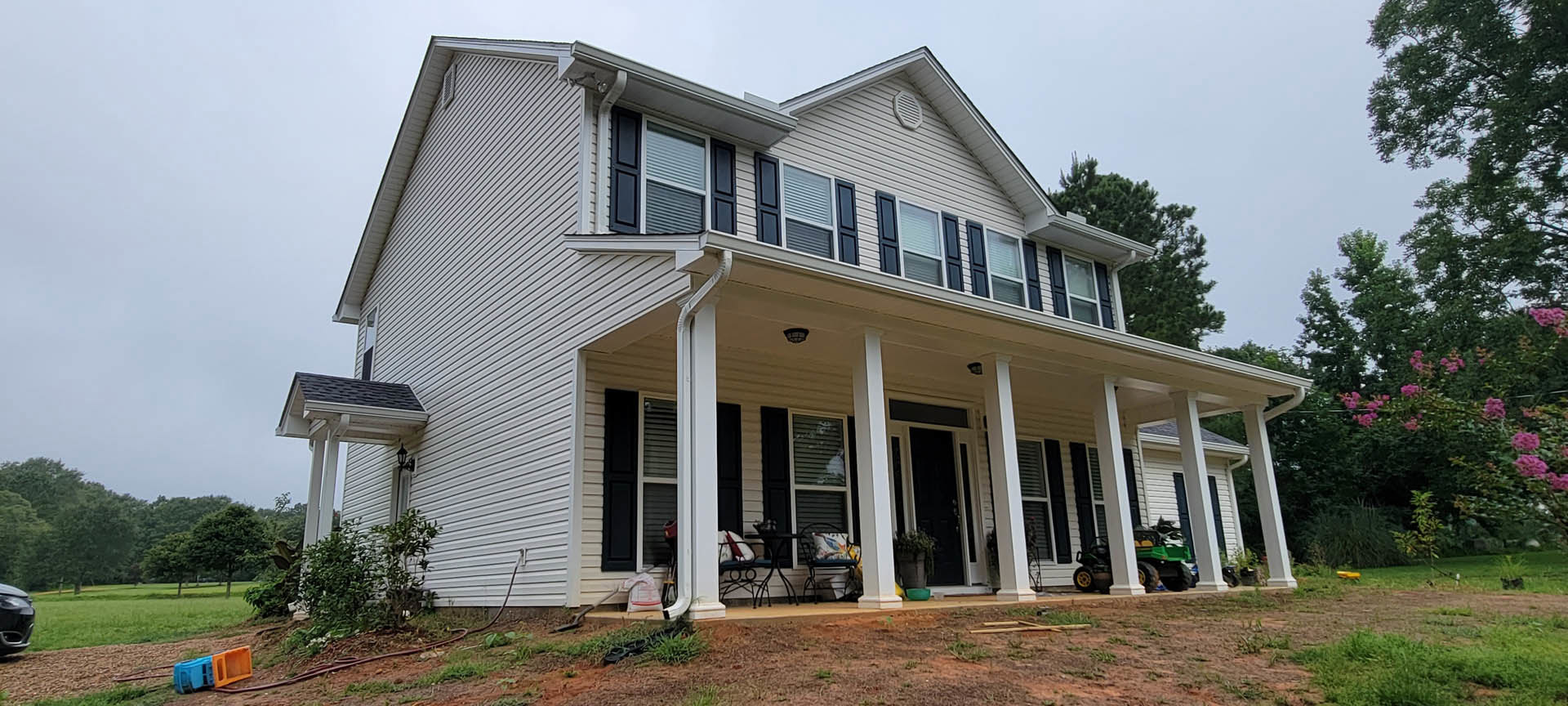 White two-story house with covered front porch, white columns, and green lawn bordered by trees; blue and orange truck and green vehicle parked in driveway.