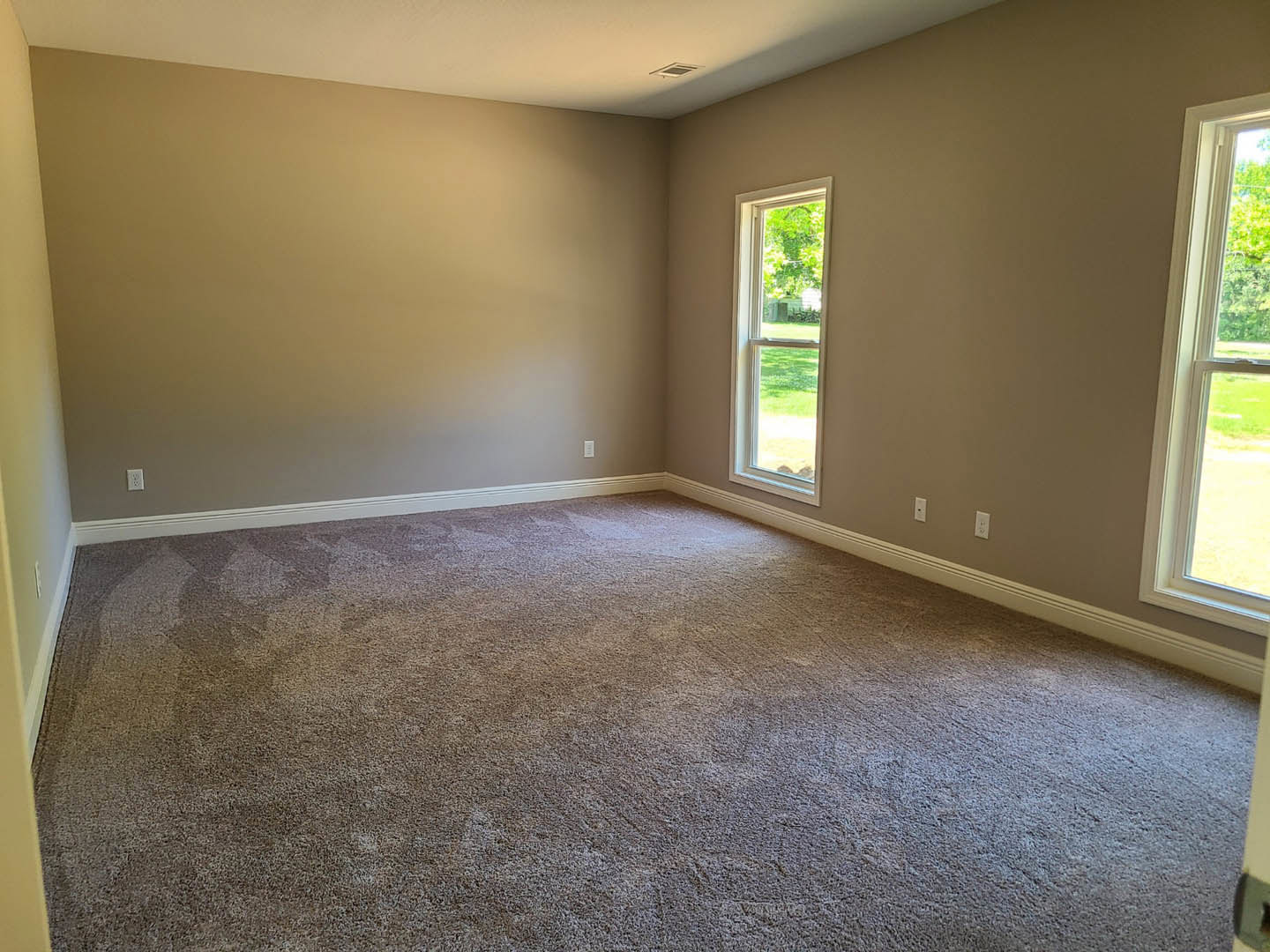 Carpeted room with large window overlooking green lawn and leafy plants, white plaster walls, and laminate flooring near the window